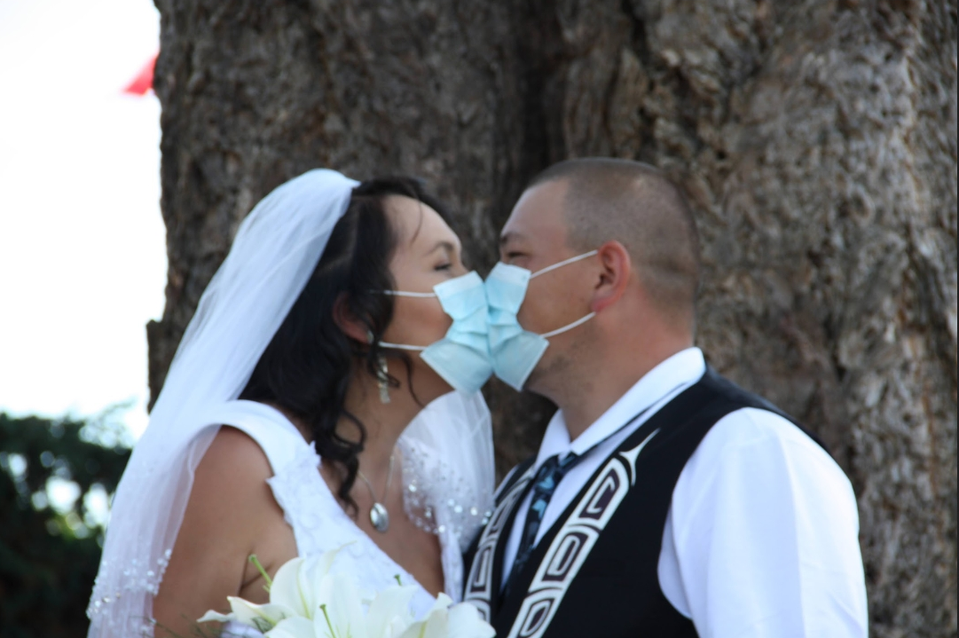 Two people dressed in wedding atire kissing through blue surgical masks