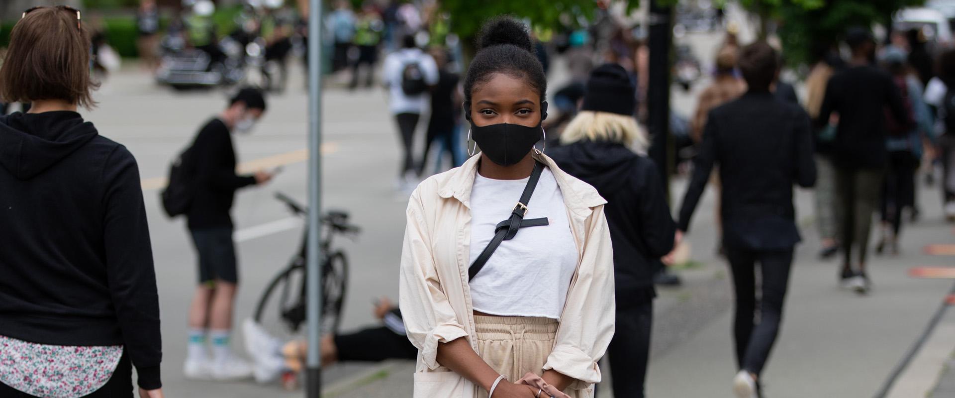 A woman wearing a black face mask standing on a busy sidewalk.