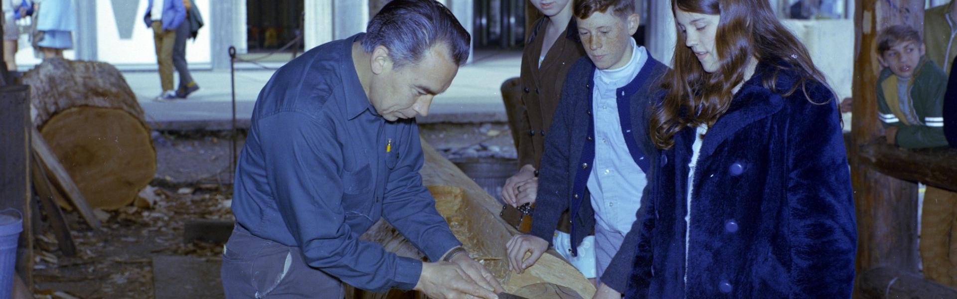 Henry Hunt carving the Mungo Martin mortuary totem, in carving studio with visitors looking on. BC Archives i-19444 