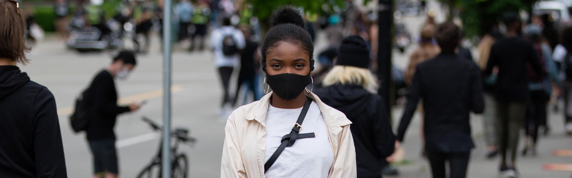 Woman standing on a busy sidewalk wearing a black face mask.
