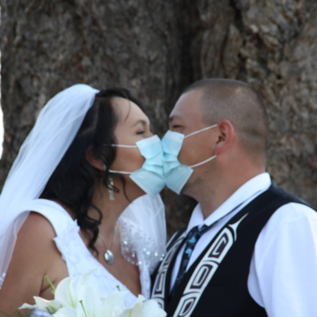 Two people dressed in wedding atire kissing through blue surgical masks