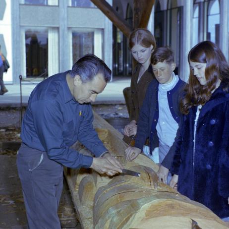 Henry Hunt carving the Mungo Martin mortuary totem, in carving studio with visitors looking on. BC Archives i-19444 